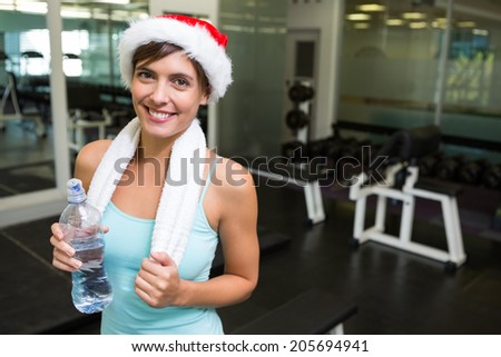 Fit brunette in santa hat smiling at camera at the gym
