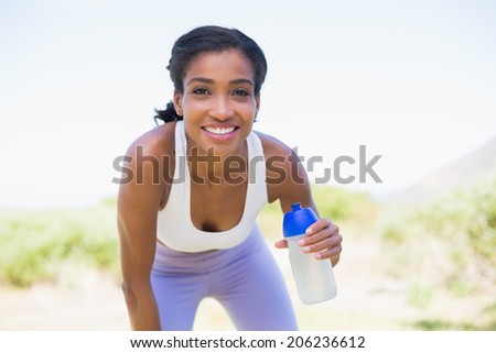 Fit woman holding sports bottle smiling at camera on a sunny day in the countryside