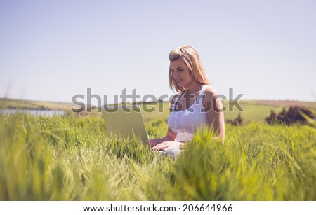 Pretty blonde sitting on grass using her laptop on a sunny day in the countryside