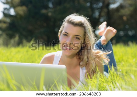 Pretty blonde lying on grass using laptop smiling at camera on a sunny day in the countryside