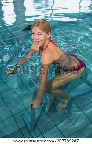 Fit blonde using underwater exercise bike smiling at camera in swimming pool at the leisure centre
