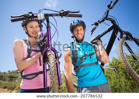 Fit couple walking down trail smiling at camera holding mountain bikes on a sunny day