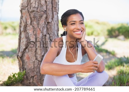 Fit woman sitting against tree listening to music on a sunny day in the countryside