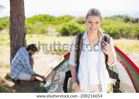 Attractive blonde smiling at camera while partner pitches tent on a sunny day