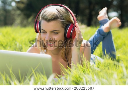 Pretty blonde lying on grass using laptop listening to music on a sunny day in the countryside