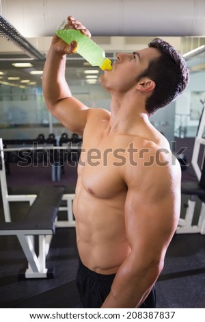 Side view of a shirtless muscular man drinking energy drink in gym
