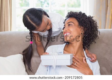 Pretty mother sitting on couch with daughter offering a gift at home in the living room