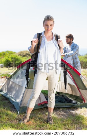 Attractive blonde smiling at camera while partner pitches tent on a sunny day