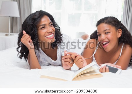 Mother and daughter reading book and listening to music together on bed at home in bedroom