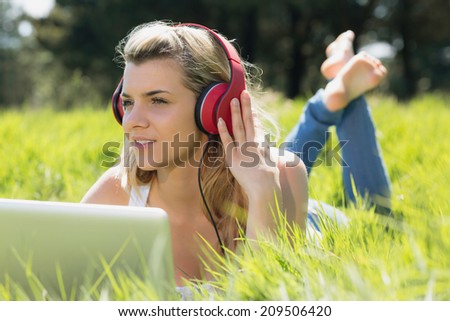 Pretty blonde lying on grass using laptop listening to music on a sunny day in the countryside