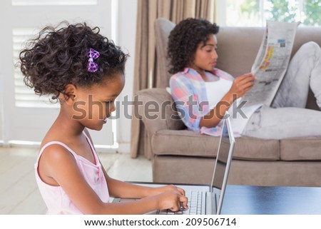 Cute daughter using laptop at desk with mother on couch at home in the living room