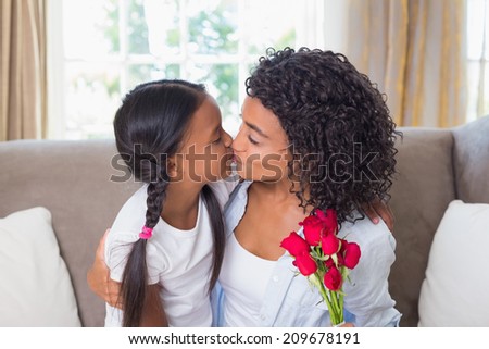 Pretty mother sitting on the couch kissing her daughter holding roses at home in the living room