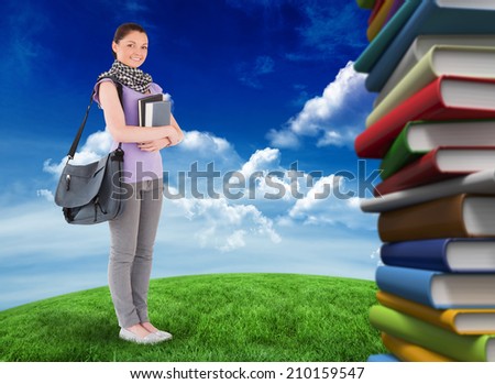 Attractive student holding books and her bag while standing against green field under blue sky