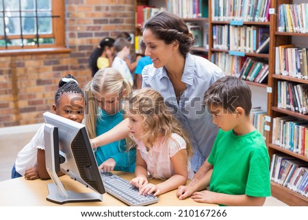 Cute pupils and teacher looking at computer in library at the elementary school