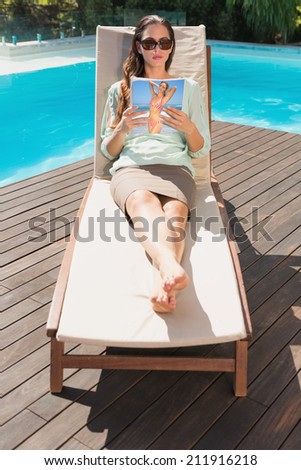 Beautiful young woman reading book on sun lounger by swimming pool