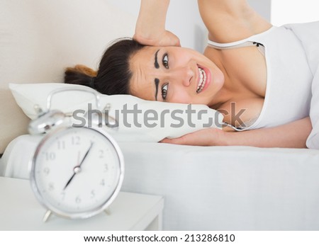 Sleepy woman covering ear with hand in bed and alarm clock on side table