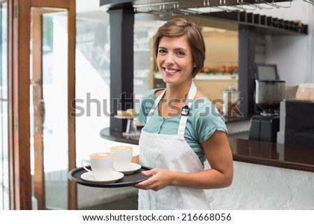 Pretty barista smiling at camera holding tray at the coffee shop
