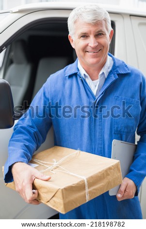 Delivery driver smiling at camera by his van holding parcel outside the warehouse