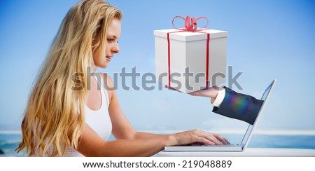 Composite image of pretty blonde using her laptop at the beach with hand holding gift