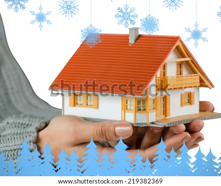 Hands showing a miniature model home against snowflakes and fir trees in blue