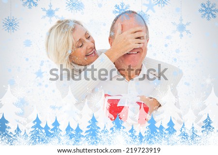 Older woman covering her partners eye while holding present against snowflakes and fir trees