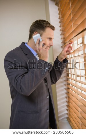 Businessman peeking through blinds while on call in office