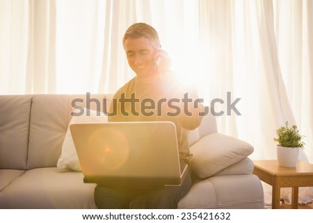 Happy man sitting on couch phoning and using laptop at home in the living room