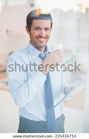 Smiling businessman holding tablet and disposable cup looking out the window in his office