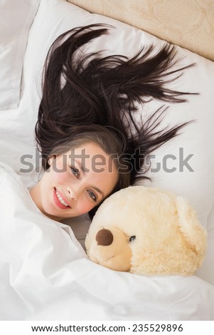 Pretty brunette under the duvet with teddy bear at home in the bedroom