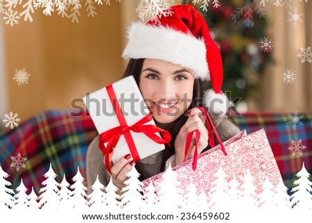Smiling brunette holding gift and shopping bag at christmas against fir tree forest and snowflakes