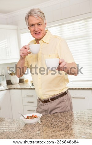 Senior man offering cup to camera at home in the kitchen