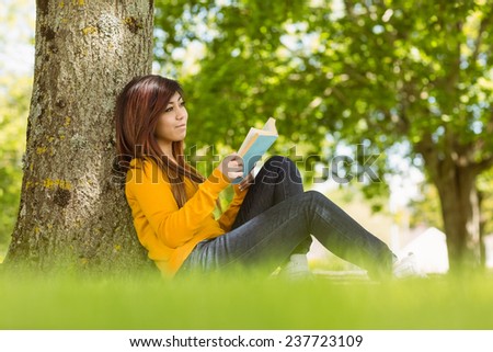 Relaxed female college student reading book against tree trunk in the park