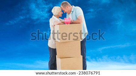 Older couple smiling at each other with moving boxes and piggy bank against blue sky