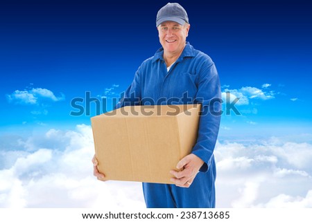 Happy delivery man holding cardboard box against bright blue sky over clouds