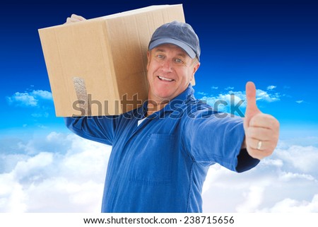 Happy delivery man holding cardboard box against bright blue sky over clouds