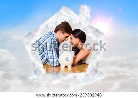 Young couple lying on floor smiling with piggy bank against blue sky with white clouds