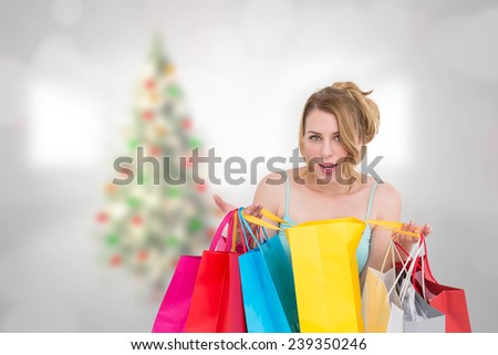Excited woman looking at camera with many shopping bags against blurry christmas tree in room