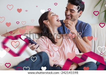 Playful couple watching TV while eating popcorn against love heart pattern