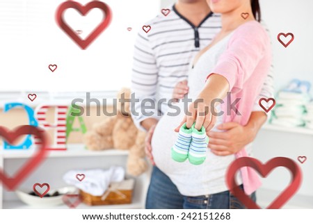 Close up of a bright pregnant woman holding baby shoes while husband touching her belly in the room against hearts