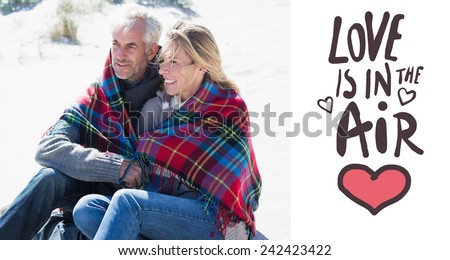 Happy couple wrapped up in blanket sitting on the beach against love is in the air