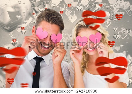 Attractive young couple holding pink hearts over eyes against grey valentines heart pattern