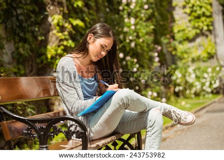 Smiling student sitting on bench and writing on notepad in park at school