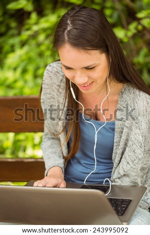 Smiling student sitting on bench listening music and using laptop in park at school