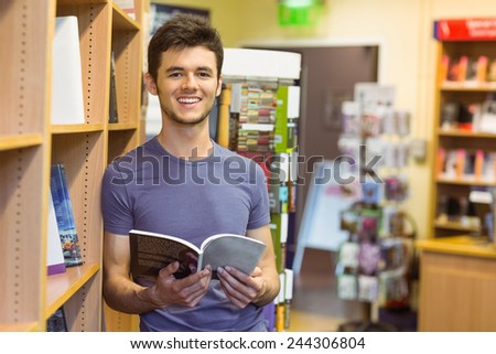 Smiling university student holding textbook in the bookcase at the university