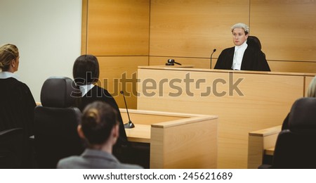Unsmiling judge wearing wig with american flag behind him in the court room