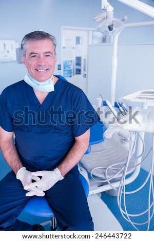 Smiling dentist in blue scrubs sitting in chair at the dental clinic