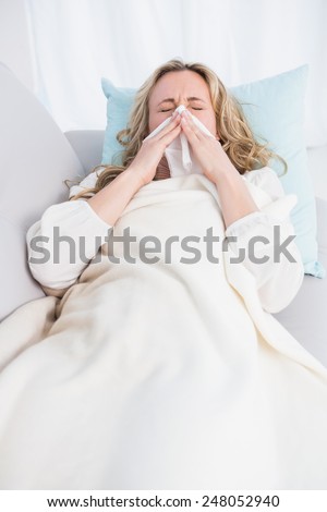 Blonde lying on couch sneezing on tissue at home in the living room