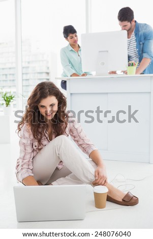 Smiling businesswoman sitting on the floor using laptop with colleagues behind her