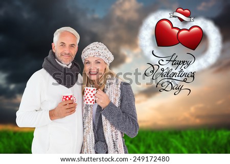 Happy couple in winter fashion holding mugs against green grass under blue and orange sky