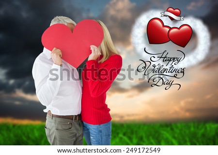 Couple embracing and holding heart over faces against green grass under blue and orange sky